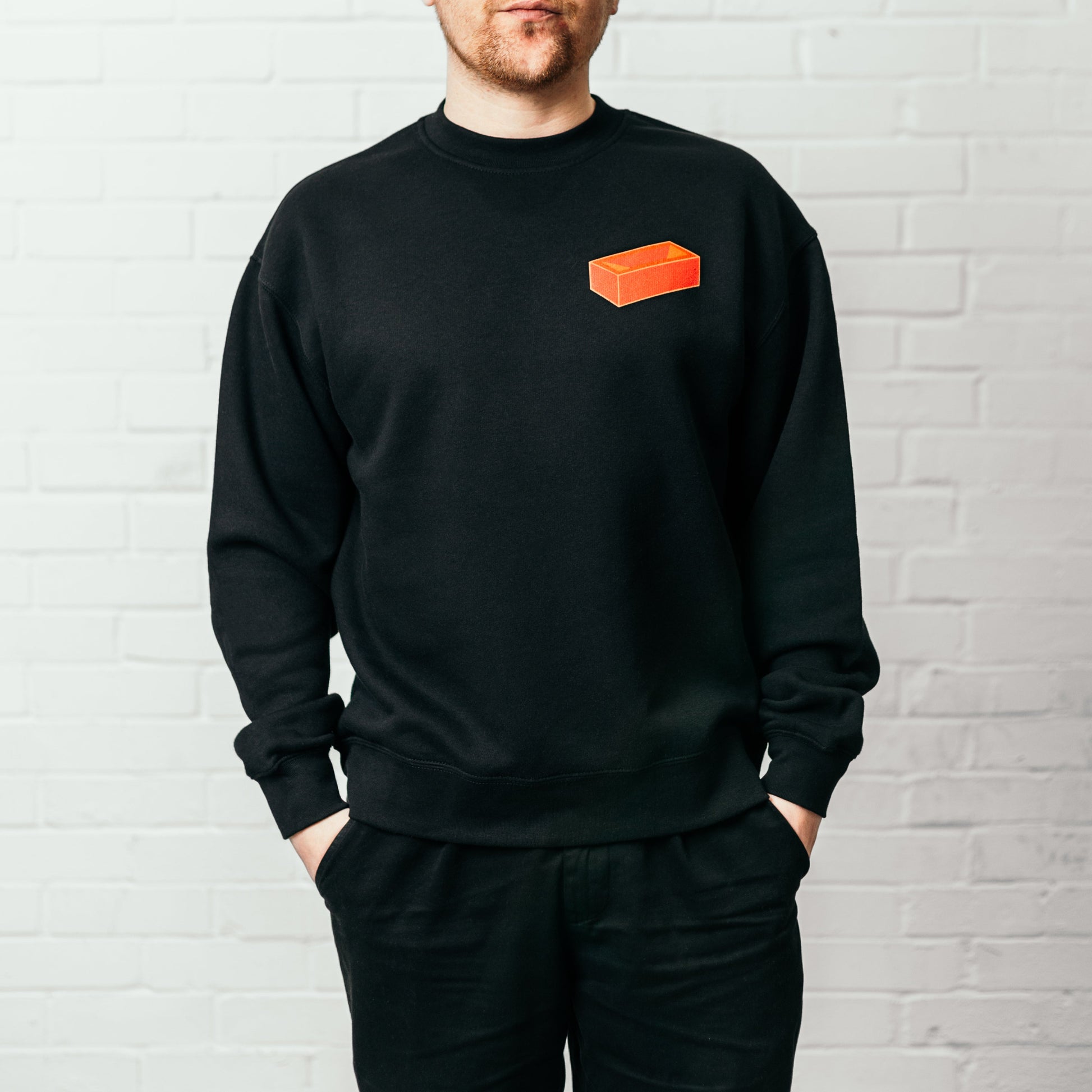 A man wearing a black crew neck sweatshirt with a red brick printed on the top left.