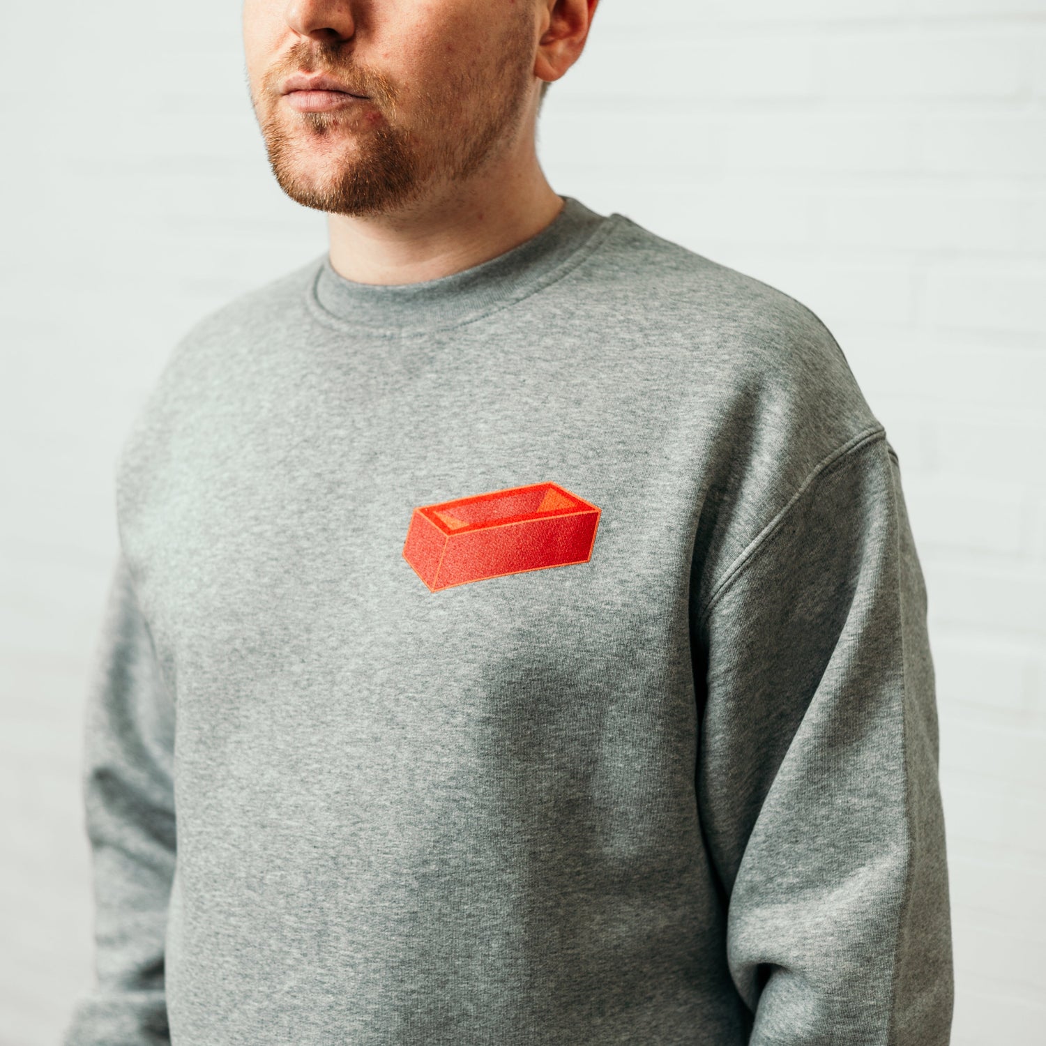 A man wearing a grey, crew neck sweatshirt featuring a printed red brick on the top left.