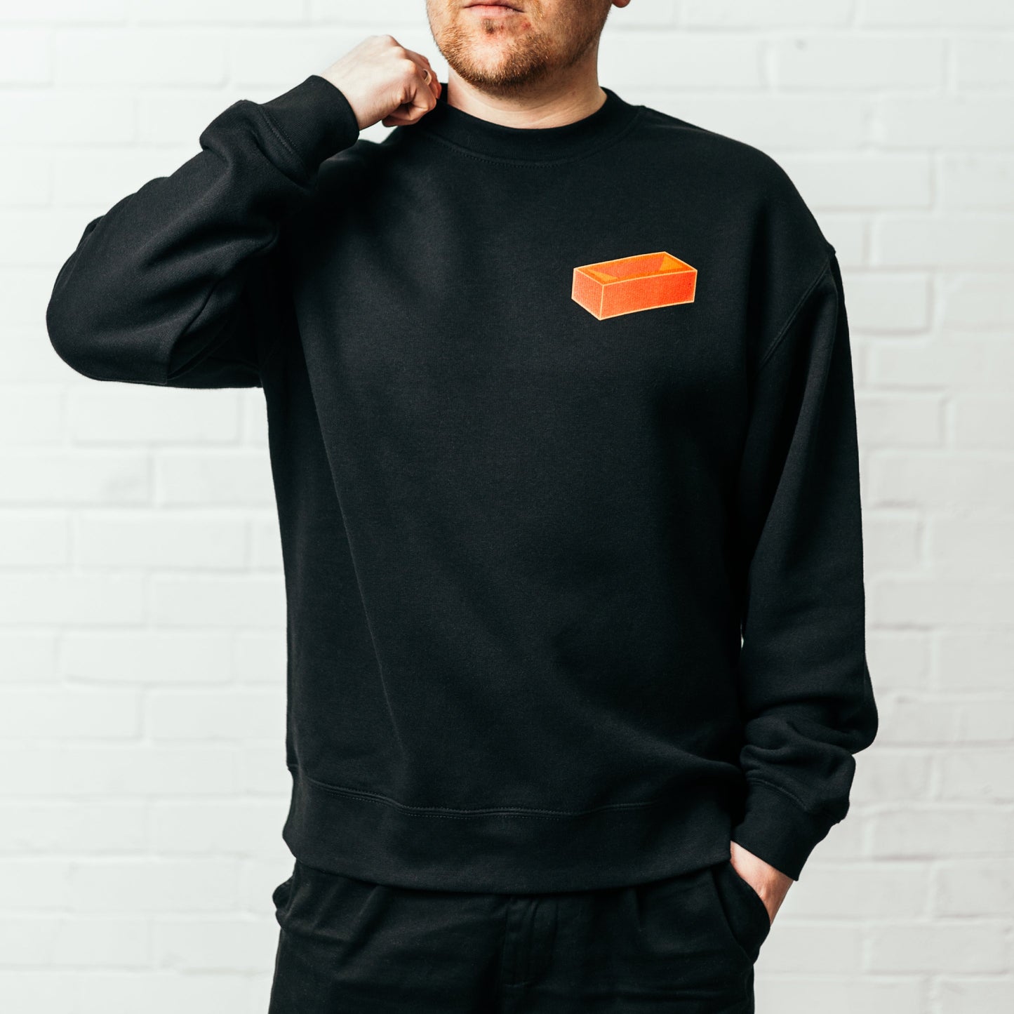 A man wearing a black crew neck sweatshirt with a red brick printed on the top left.